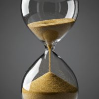 A close-up shot of a transparent hourglass with golden sand flowing between its two bulbs, symbolizing the passage of time and deadlines. The dark, neutral background adds a sense of reflection and