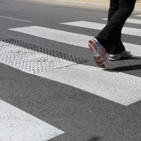 A person wearing athletic shoes crosses a sunlit pedestrian crosswalk on an asphalt road. The scene conveys movement and urban life.
