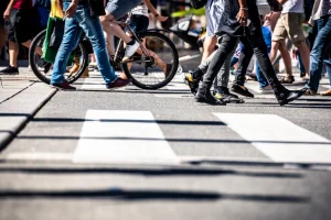 A bustling city scene shows a diverse crowd crossing a zebra crossing, with a cyclist among them. Sunlight creates long shadows, conveying a lively urban vibe.