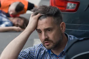 A distressed man in a checkered shirt sits by a car, holding his head in his hand, while an officer talks to him. Two injured people lie on the road in the background.