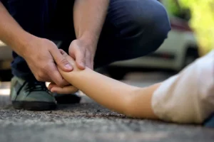 A person crouches on a road, gently checking the pulse of an individual lying down, with a car in the background, conveying urgency and care.
