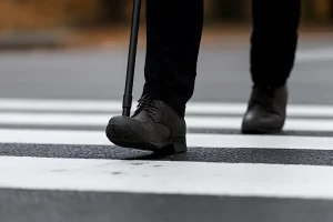 A person in black pants and boots uses a cane to walk across a zebra crosswalk. The scene suggests caution and focus on mobility.