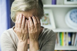 An elderly person with short gray hair covers their face with both hands, conveying stress or sadness. A blurred bookshelf and clock are in the background.